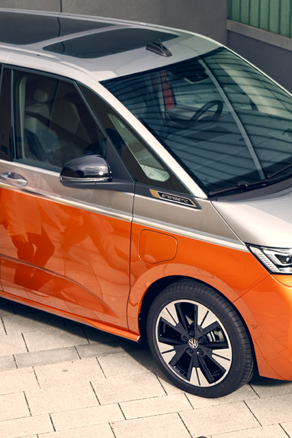 Volkswagen Multivan – перфектен за семейни приключения. Щастливо семейство с деца пред оранжев Volkswagen Multivan, паркиран до сграда с електрическо зарядно.