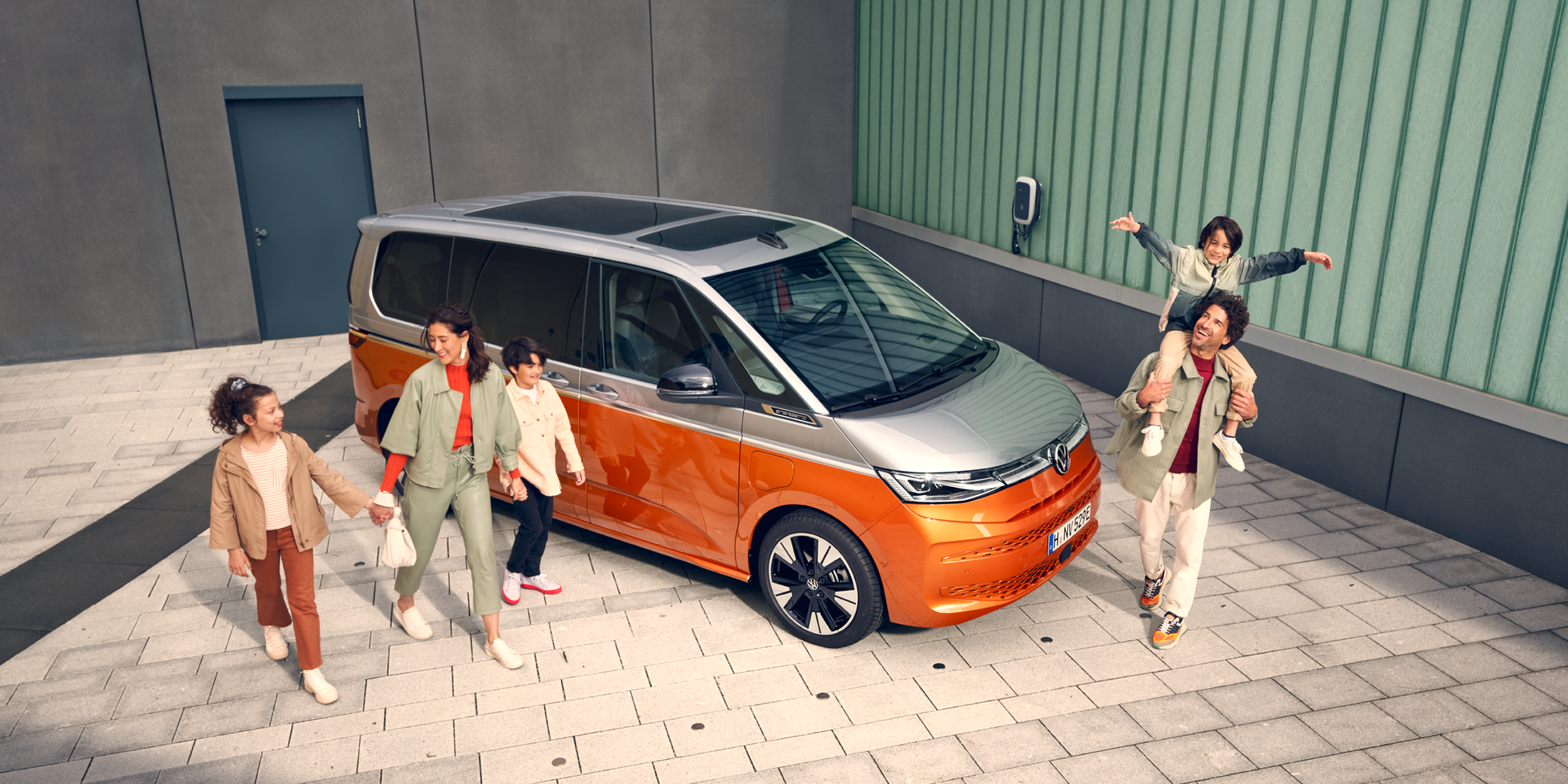 Volkswagen Multivan – перфектен за семейни приключения. Щастливо семейство с деца пред оранжев Volkswagen Multivan, паркиран до сграда с електрическо зарядно.